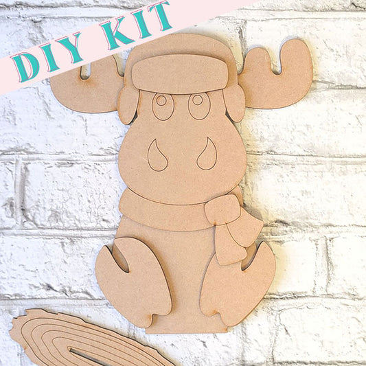 Introducing the Woodland Friends Moose Shelf Sitter from Janet's Craft Corner: a DIY kit featuring precut cardboard pieces to assemble a charming moose adorned with a scarf and hat. Ideal for home decor, this delightful project comes against a light brick background.