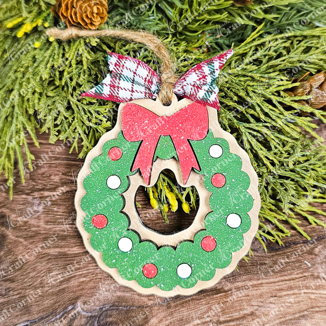 A wooden ornament from Janet's Craft Corner, part of the Sugar Cookie Ornaments - Set of 3, captures the delightful appearance of a sugar cookie. The wreath shimmers with green glitter, red and white circles, and is topped with a ribbon in red, white, and green plaid. The ornament hangs gracefully against a backdrop of wood and lush green foliage.