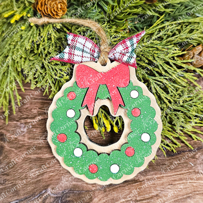 A wooden ornament from Janet's Craft Corner, part of the Sugar Cookie Ornaments - Set of 3, captures the delightful appearance of a sugar cookie. The wreath shimmers with green glitter, red and white circles, and is topped with a ribbon in red, white, and green plaid. The ornament hangs gracefully against a backdrop of wood and lush green foliage.