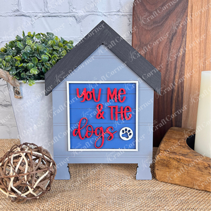 The Dog Squares by Janet's Craft Corner is a blue and gray doghouse-shaped sign with red and white "You, Me & The Dogs" text and a paw print—perfect for tiered tray decor, shown on a woven mat with plants and wood accents.