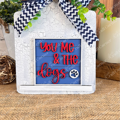 The Dog Squares sign by Janet's Craft Corner features a rustic white frame, blue background, red "You Me & The dogs" text with a paw print, chevron bow, faux greenery, and sits on a woven mat—ideal for tiered tray decor.