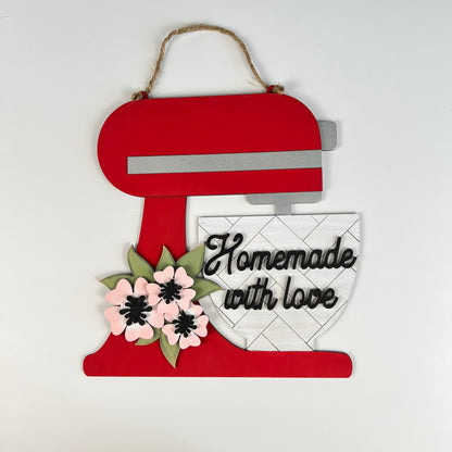 The Kitchen Mixer Wall Hanger by Janet's Craft Corner is a delightful DIY kit that includes a decorative wall piece shaped like a red kitchen mixer. It features vibrant pink and black flowers and the heartwarming phrase "Homemade with love" on the mixing bowl. This charming piece of home decor can be easily hung using twine.