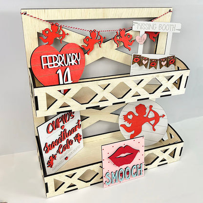 A charming display from Janet's Craft Corner's Cupid Kissing Booth Tiered Tray Kit showcases delightful Valentine's Day decor, featuring a "February 14" heart, cherub cutouts, a playful "Cupid Kissing Booth" sign, and a whimsical "Smooch" sign with lips. The tiered tray boasts a geometric cut-out design ideal for any DIY kit enthusiast.