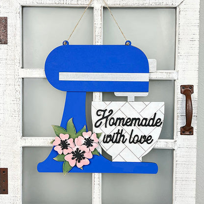 A captivating wall hanger from Janet's Craft Corner, the Kitchen Mixer Wall Hanger is designed to resemble a blue stand mixer complete with a bowl. Embellished with the words "Homemade with love" and adorned with pink flowers, this charming piece of home decor hangs elegantly on a white paneled door, featuring a rustic handle.