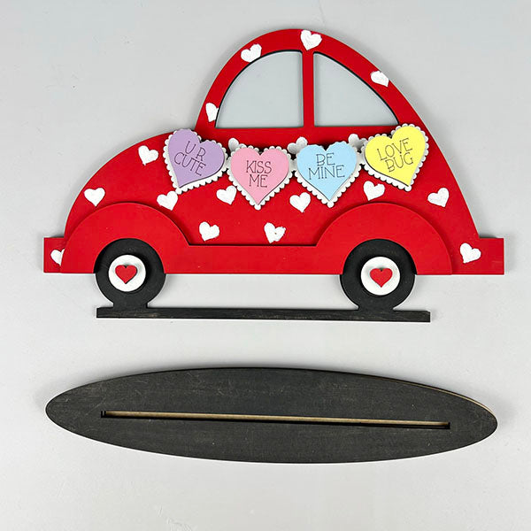 The Valentine Volkswagen Shelf Sitter by Janet's Craft Corner is a delightful red car-shaped decoration adorned with small white hearts and features four heart-shaped messages: "UR CUTE," "KISS ME," "BE MINE," and "LOVE BUG." It gracefully sits on a black oval base with a handy slot, ideal for displaying your handmade crafts.