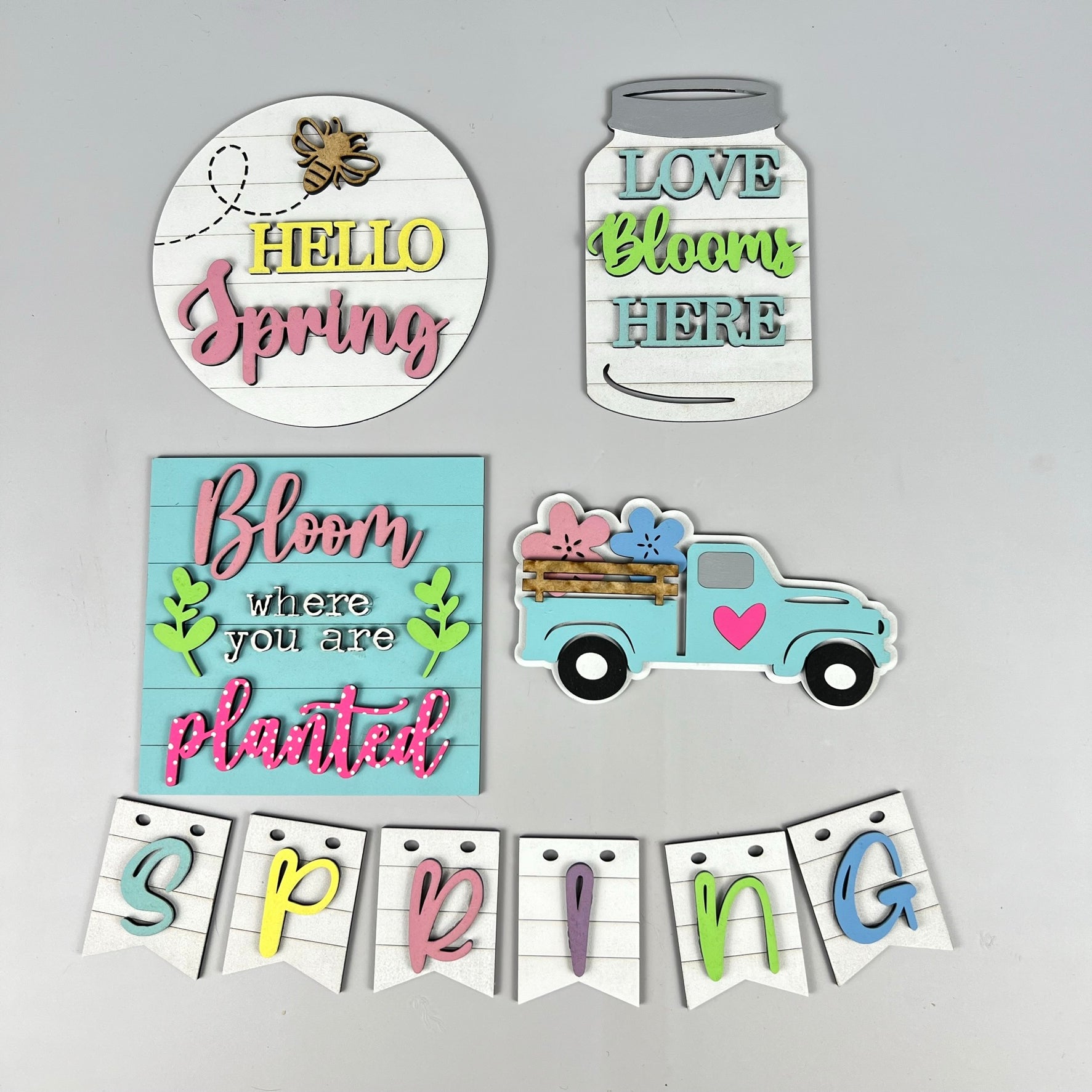 Introducing the Spring Truck Tiered Tray Kit from Janet's Craft Corner, featuring decorative spring-themed wooden signs and cutouts. This kit is perfect for your tiered tray and includes a bee with "Hello Spring," a mason jar with "Love Blooms Here," a truck loaded with flowers, charming phrases like "Bloom where you are planted," and a cheerful "SPRING" banner.