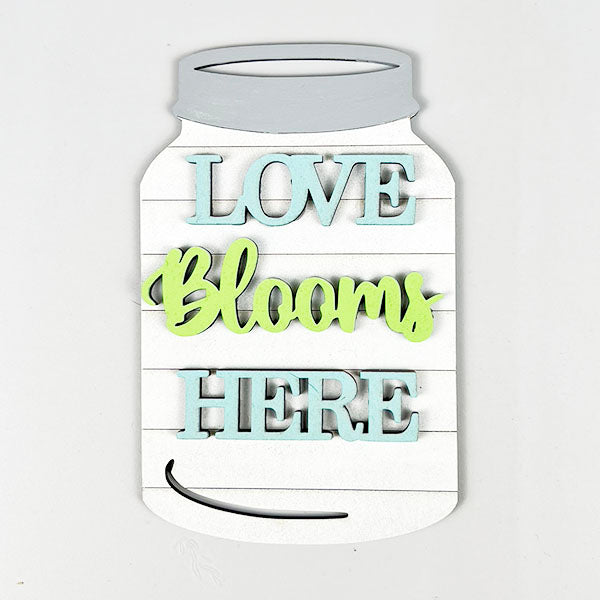 Add a charming touch to your spring decor with Janet's Craft Corner's Spring Truck Tiered Tray Kit. This DIY project features a mason jar-shaped decorative sign boldly displaying "Love Blooms Here" in colorful letters, with "Love" and "Here" in blue and "Blooms" in green against a background that mimics white wooden planks. Perfect for enhancing your tiered tray kit display!