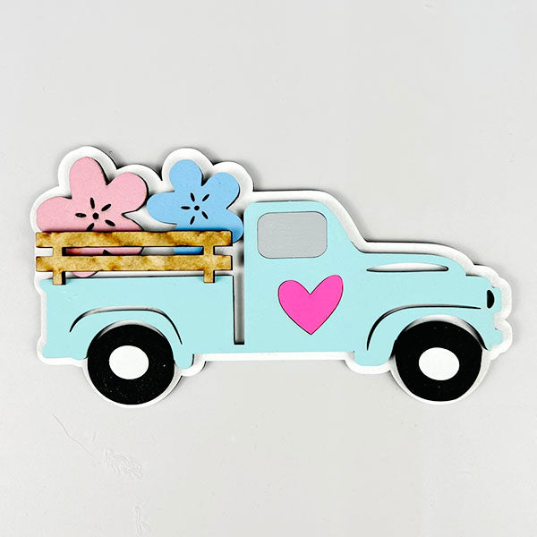 Illustration of a light blue vintage truck with a pink heart on its side, carrying large pink and blue flowers in the back. This Spring Truck Tiered Tray Kit from Janet's Craft Corner features wooden rails and black wheels, set against a plain white background—ideal for your spring decor needs.