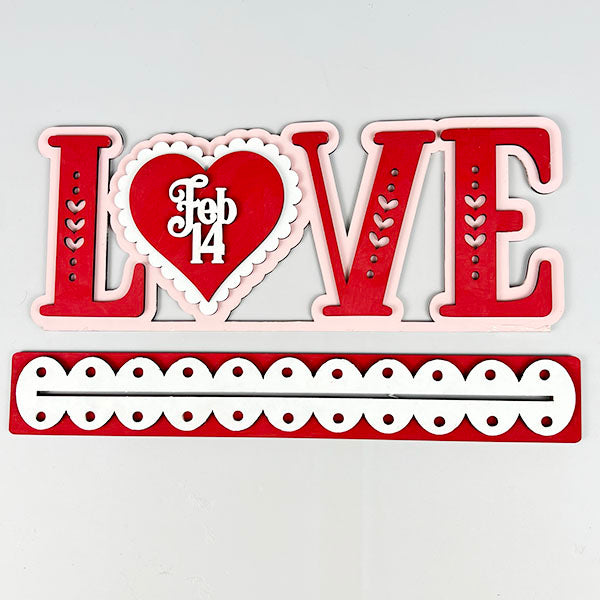 The "Love Shelf Sitter" from Janet's Craft Corner is a delightful home décor piece that showcases the word “LOVE” in red and pink, with a heart-shaped “O” and “Feb 14” in white. It features a charming red and white border adorned with circular cutouts, making it perfect for craft kits or DIY kit enthusiasts looking to personalize their space.