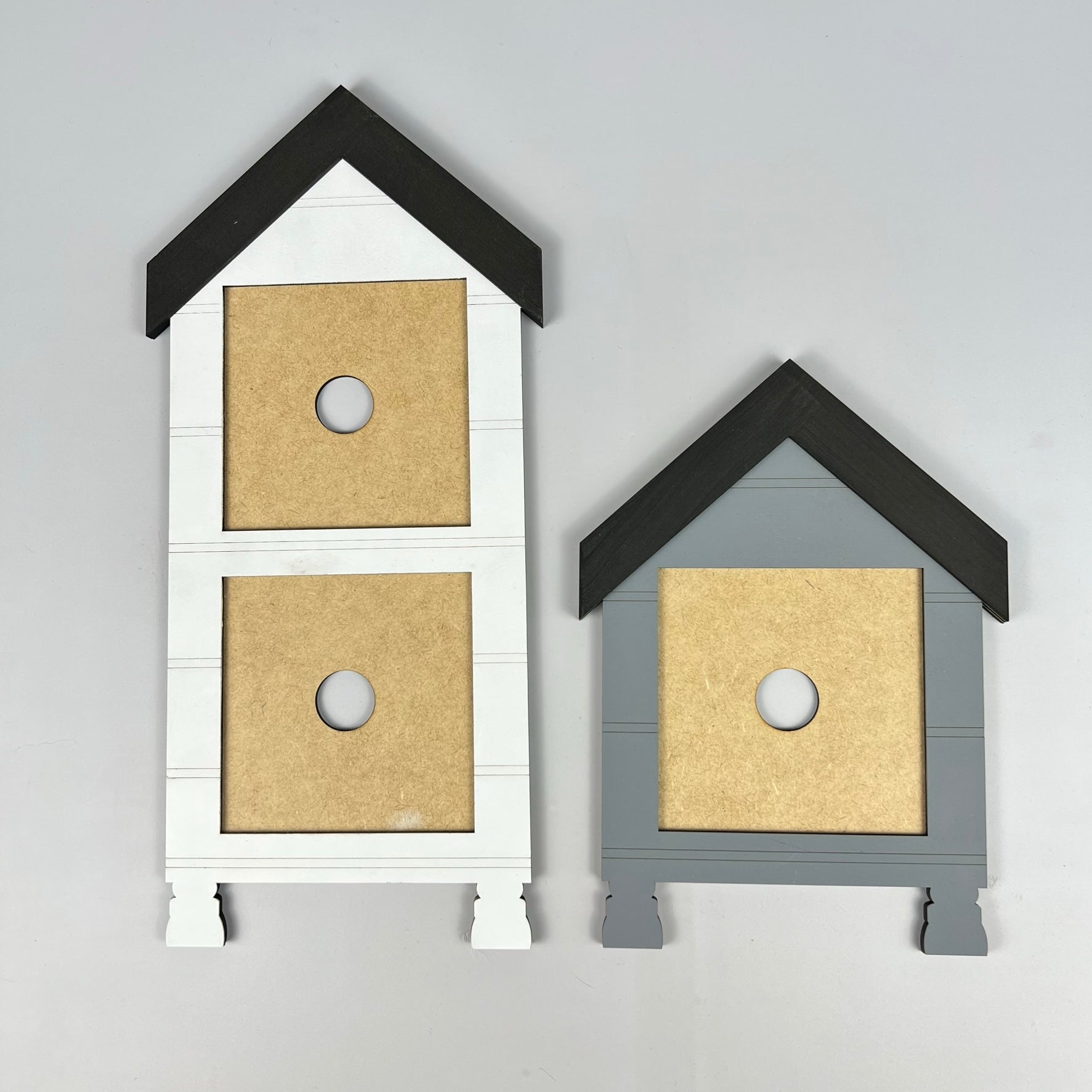 On a gray backdrop, the House Interchangeable Squares Holders from Janet's Craft Corner showcase two charming wooden birdhouse fronts that evoke farmhouse décor. The taller one is painted white with two circular openings, while the shorter gray one features just one opening. Both designs boast dark roofs and natural wood sections, making them perfect for your next DIY kit or seasonal craft project.