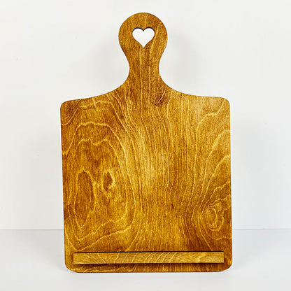 Introducing the Breadboard Interchangeable Squares Holder by Janet's Craft Corner: a delightful wooden kitchen stand with a heart-shaped cutout at the top, ideal for supporting tablets or cookbooks. It boasts a natural wood grain finish and includes a small ledge at the bottom, making it perfect as both a breadboard sign holder for interchangeable square signs in your DIY kit projects.