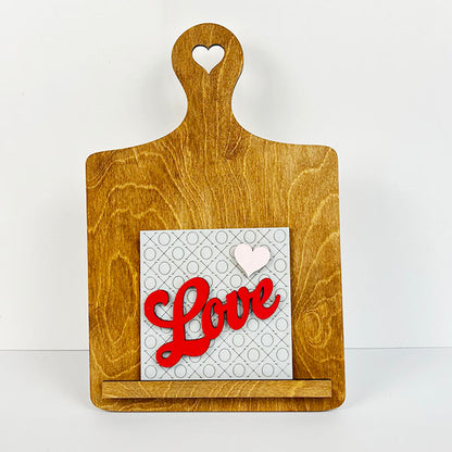 A Breadboard Interchangeable Squares Holder from Janet's Craft Corner, featuring a heart-shaped cutout in the handle, serves as a delightful sign holder. It displays a card with "Love" written in red script alongside a white heart on a patterned background. The board stands elegantly against a white backdrop, ready to inspire your next DIY kit project.
