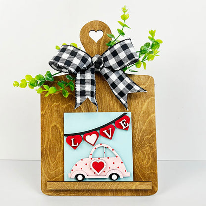 Introducing the Breadboard Interchangeable Squares Holder from Janet's Craft Corner. This DIY kit includes a wooden cutting board adorned with a black and white checkered ribbon and green leaves. It features a unique interchangeable square sign, showcasing a pink car embellished with hearts under a "LOVE" banner against a blue background—ideal as a decorative breadboard sign holder.