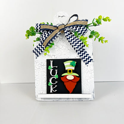 A charming Breadboard Interchangeable Squares Holder from Janet's Craft Corner showcases a white, textured plaque adorned with green foliage and a black and white zigzag bow. It includes a delightful St. Patrick's Day gnome with a top hat and beard beside the word "LUCK," making it perfect for displaying interchangeable square signs.