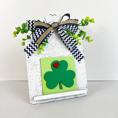 From Janet's Craft Corner, the Breadboard Interchangeable Squares Holder features a charming three-leaf clover and a small red heart on a green square. It's elegantly adorned with chevron-patterned ribbon and lush foliage on a textured white board, highlighted by a delightful heart cut-out at the top.