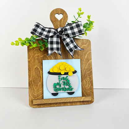 Presenting the delightful Breadboard Interchangeable Squares Holder by Janet's Craft Corner, featuring a charming wooden design adorned with a checkered bow and green foliage. This DIY kit includes an interchangeable square sign that vividly showcases a pot of gold alongside the bold phrase "End of the Rainbow" in striking green.
