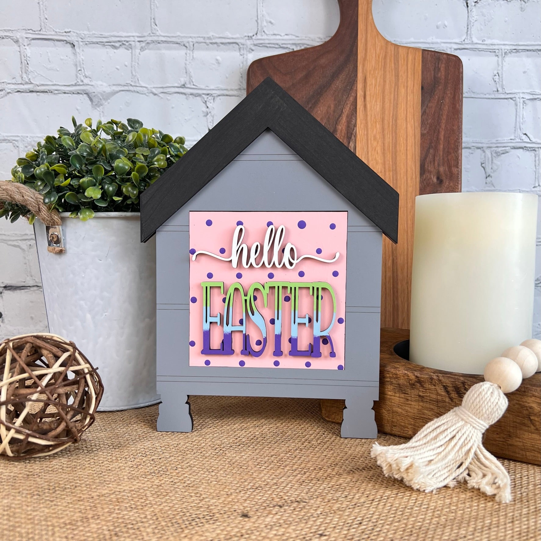 Add a touch of farmhouse décor with the House Interchangeable Squares Holders from Janet's Craft Corner. This decorative item, featuring "hello EASTER" in colorful letters, pairs beautifully on a table with a small potted plant, a wooden cutting board, and a large white candle—ideal for seasonal craft enthusiasts.