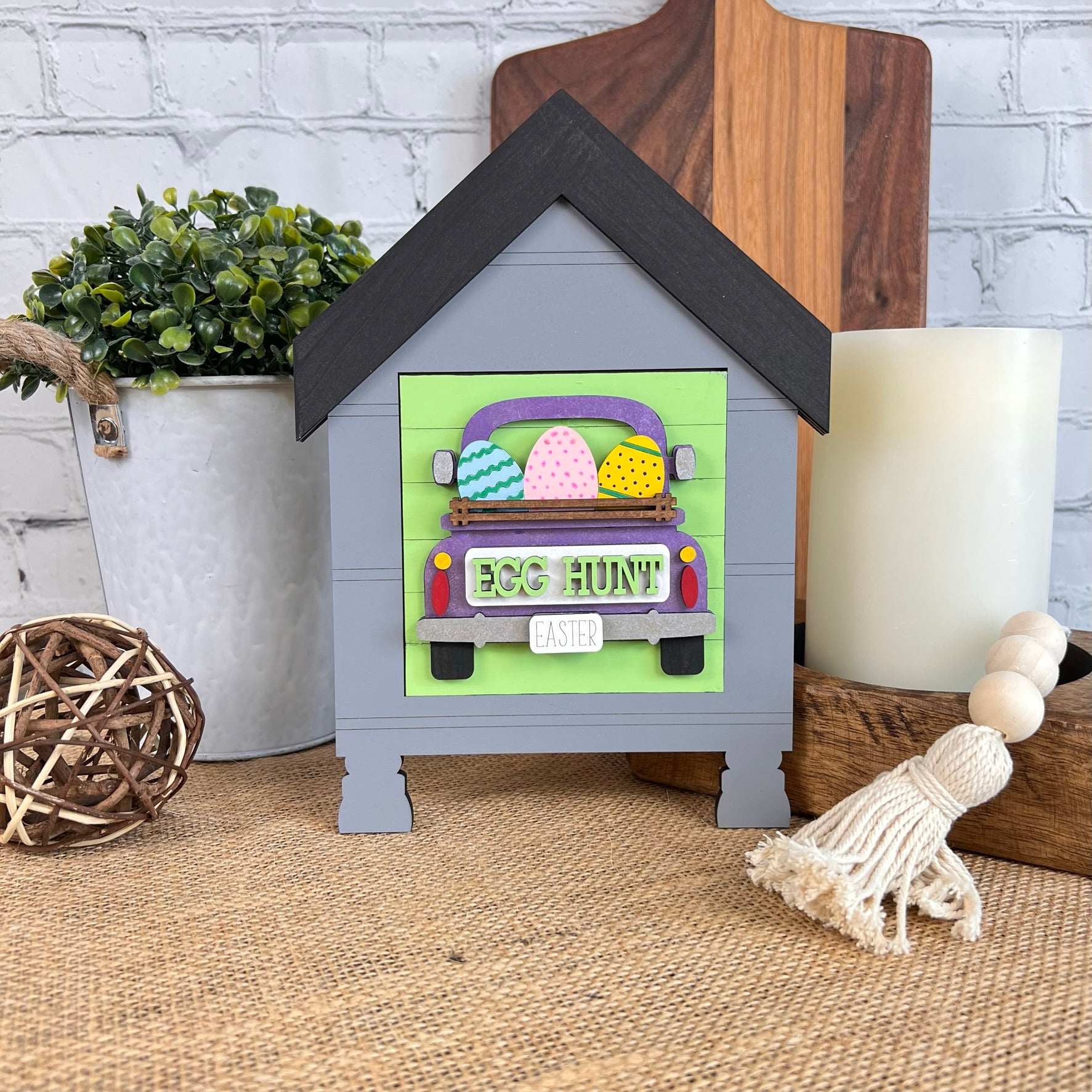 The House Interchangeable Squares Holders by Janet's Craft Corner presents a charming Easter-themed display with a wooden house shape and a truck carrying an assortment of colorful eggs, complete with "Egg Hunt" inscribed on the bumper. This craft kit is ideal for DIY enthusiasts and is set against a textured surface, complemented by a candle, tassel, and plant in the background.