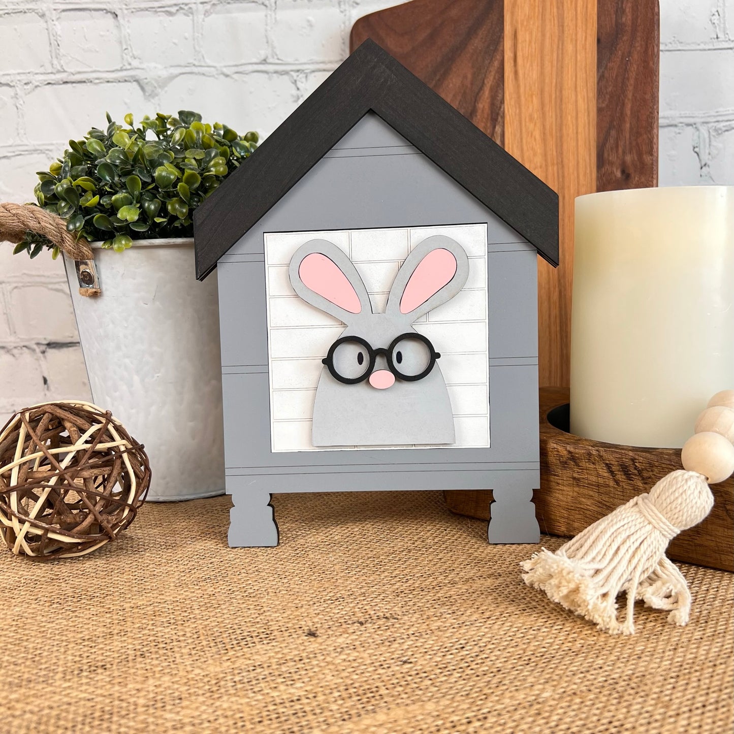 This charming item from Janet's Craft Corner, known as the House Interchangeable Squares Holders, showcases a bunny with glasses nestled within a wooden house-shaped frame. It is beautifully accented with a small plant, an illuminated candle, a woven ball, and a tassel on burlap against a white brick backdrop for that perfect farmhouse décor touch.