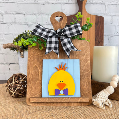 A Breadboard Interchangeable Squares Holder by Janet's Craft Corner showcases a vibrant illustration of a chick with orange feathers and a purple bow tie against a blue background. Complementary decor includes greenery, a candle, and rustic elements for extra charm.