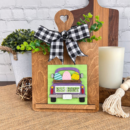 The Breadboard Interchangeable Squares Holder from Janet's Craft Corner includes a decorative wooden board featuring a green truck design with colorful Easter eggs in the trunk and "Egg Hunt!" written on it. It is adorned with a black-and-white checkered bow and fits seamlessly into an interchangeable square sign holder, making it ideal for pairing with plants and rustic decor elements.