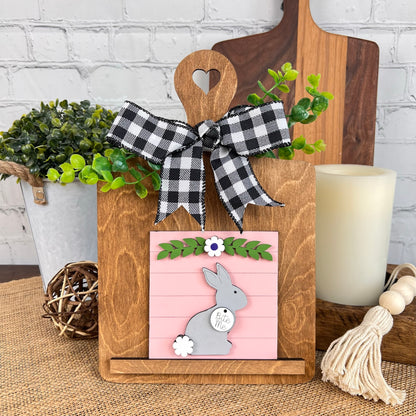 Introducing the Breadboard Interchangeable Squares Holder from Janet's Craft Corner: a charming wooden cutting board featuring a heart-shaped handle and decorated with a black-and-white checkered bow. This unique piece includes a pink panel displaying an adorable gray bunny among green leaves and white flowers. Ideal either as an interchangeable square sign or as part of your DIY projects, it pairs wonderfully with nearby plants and candles for an inviting display.