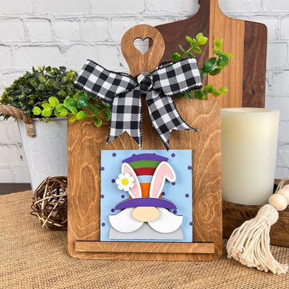 Introducing the Breadboard Interchangeable Squares Holder by Janet's Craft Corner. This decorative wooden stand features a vibrant Easter bunny sign with a colorful hat and ears. Topping it off is a black and white checkered bow, perfectly capturing the charm of an interchangeable square design. Pair it with greenery, candles, and rustic décor to create a delightful display.