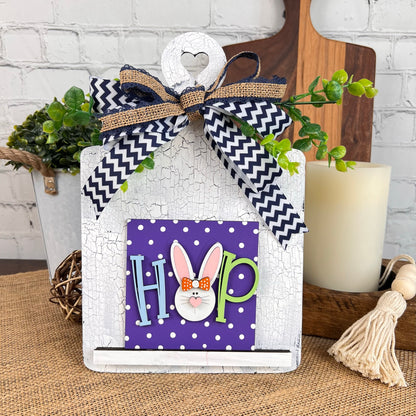 Introducing the Breadboard Interchangeable Squares Holder from Janet's Craft Corner: This charming decorative wooden board serves as both a breadboard and sign holder. It features a "HOP" sign where the "O" is creatively designed with a cartoon bunny face, complete with a polka dot bow. The piece is elegantly tied with a chevron-patterned ribbon and enhanced by greenery accents, all set against a rustic backdrop featuring candlelight and burlap for added warmth.