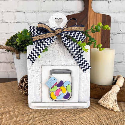 The Breadboard Interchangeable Squares Holder by Janet's Craft Corner is a decorative wooden sign holder featuring a heart cutout at the top and an illustration of a jar filled with colorful jelly beans. It includes an interchangeable square sign that hangs from a black-and-white chevron ribbon. With its rustic background, it's perfect for DIY enthusiasts who enjoy incorporating candles and plants into their decor.