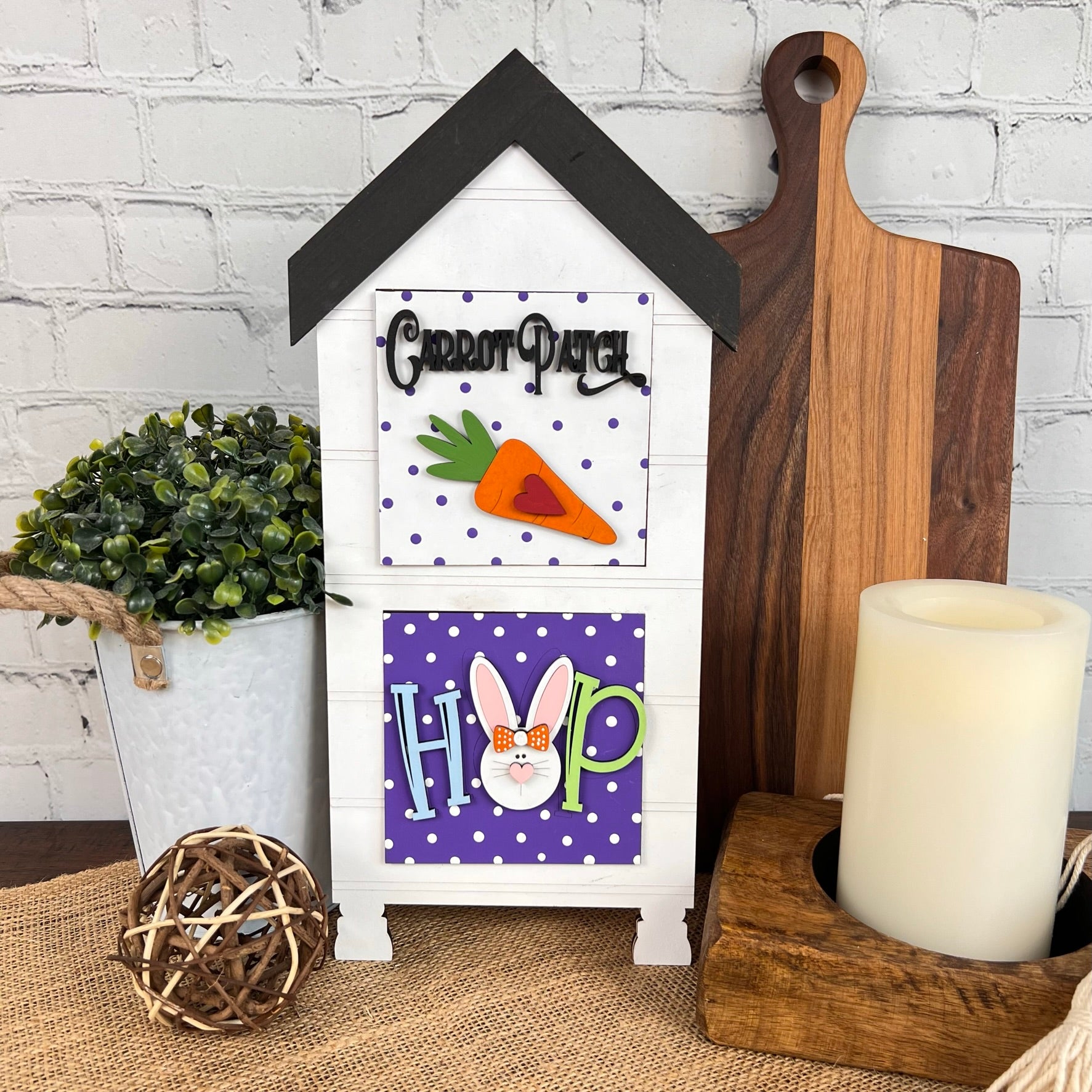 This "House Interchangeable Squares Holders" from Janet's Craft Corner, with its wooden house-shaped design featuring a "Carrot Patch" sign and a bunny face, beautifully complements your table setting. Pair it with a potted plant and candle to achieve the perfect seasonal craft kit ambiance against the white brick wall.