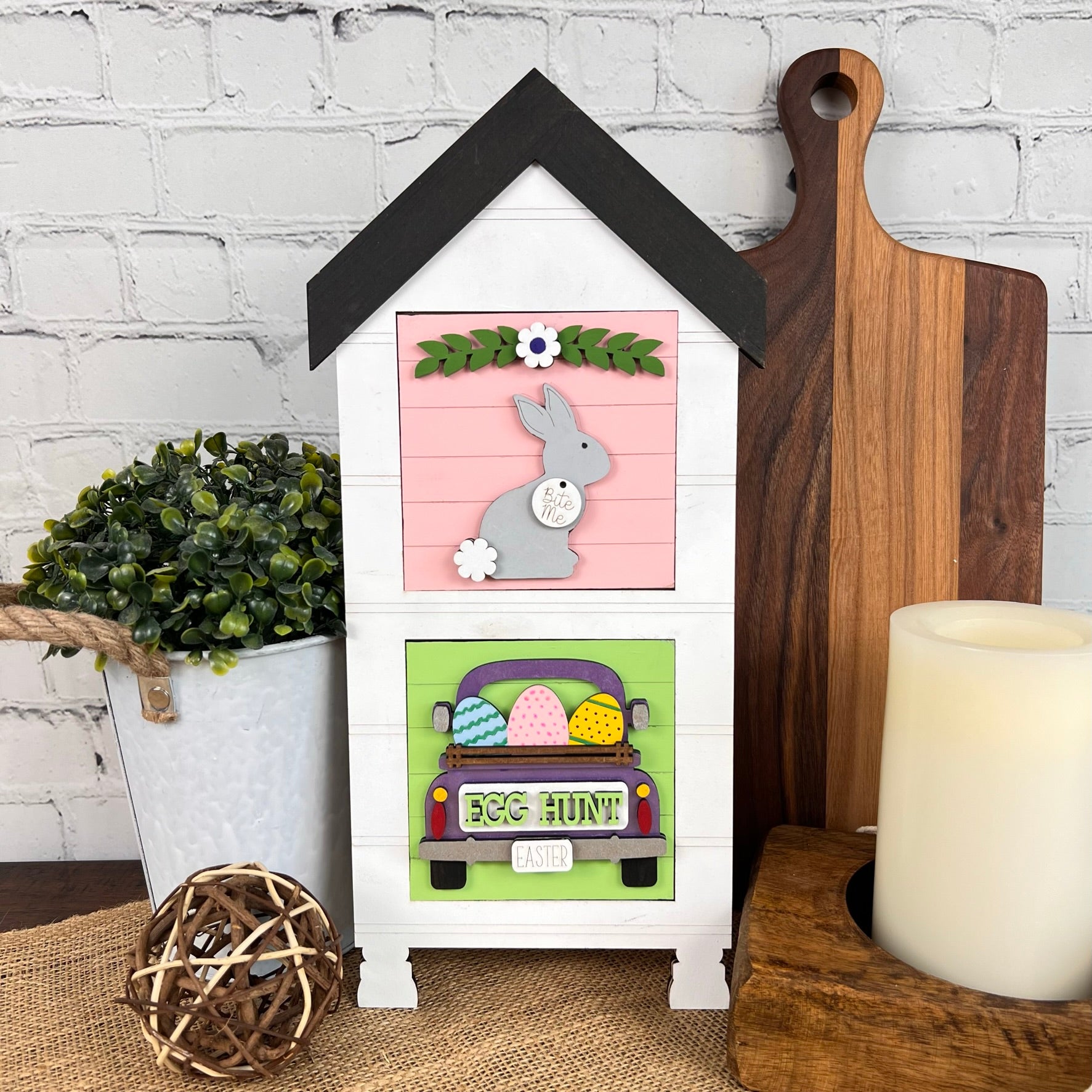 The House Interchangeable Squares Holders by Janet's Craft Corner is a delightful decorative piece that features a gray bunny and flowers with a "Happy Easter" sign, complete with an egg-filled truck. This charming farmhouse décor is beautifully set against a brick wall, alongside a plant, wooden boards, and a candle, making it an ideal addition to any seasonal craft kit collection.