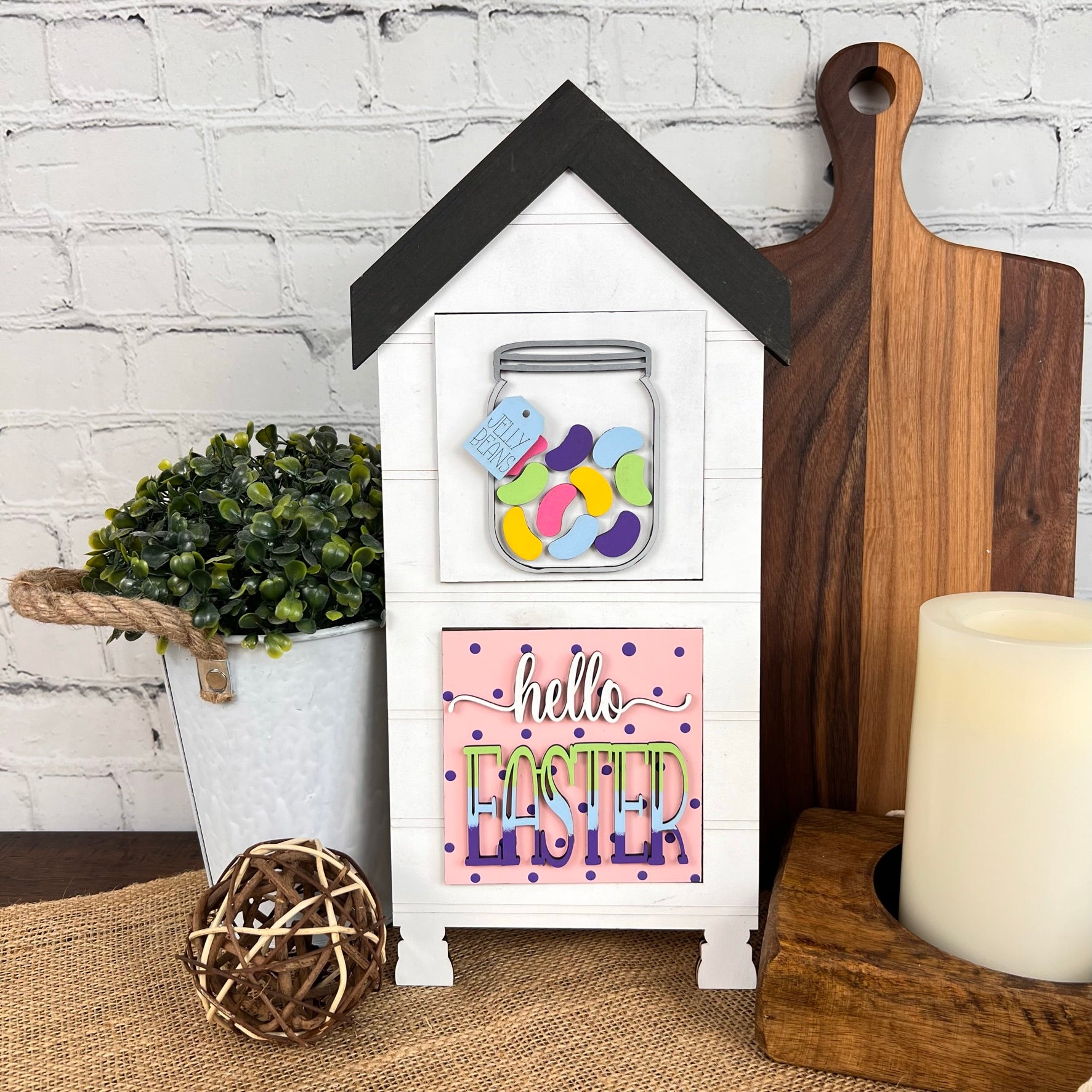 A decorative wooden House Interchangeable Squares Holder by Janet's Craft Corner, embodying farmhouse décor, showcases a jar of vibrant jelly beans and a "hello EASTER" sign. It sits on a table adorned with a potted plant, wooden cutting boards, a wicker ball, and a candle against an inviting brick wall backdrop.