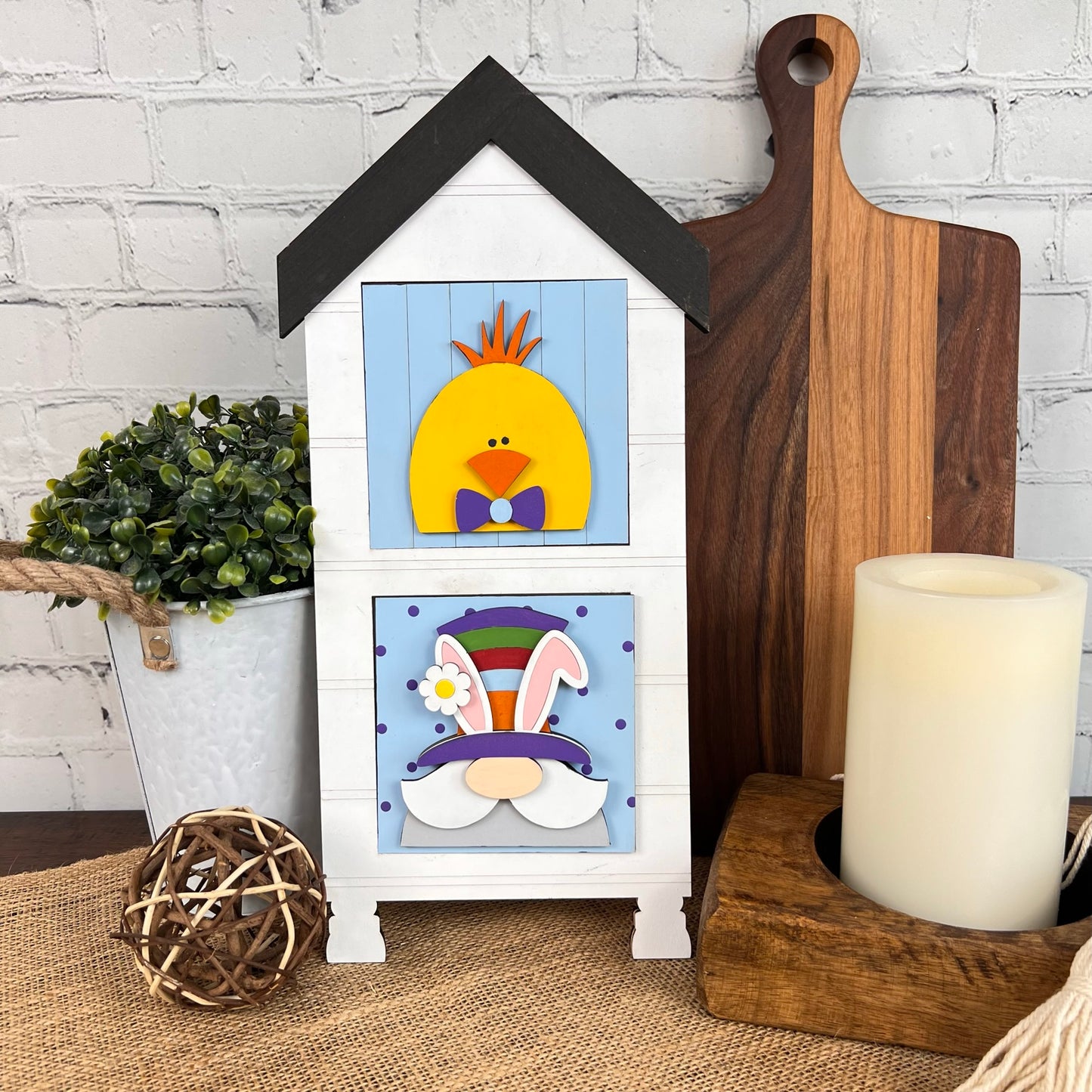 The scene features an enchanting decorative display with Janet's Craft Corner's "House Interchangeable Squares Holders," a white house-shaped piece embellished with charming chick and rabbit motifs on its doors. Embodying farmhouse style, it is complemented by a potted plant, a wooden board, a candle, and a woven ball set against a white brick background.