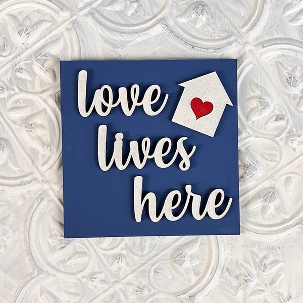 Decorative "Home Family Interchangeable Squares" from Janet's Craft Corner features a charming sign on a textured background that reads "love lives here." It includes a house-shaped cutout with a red heart, perfect for adding elegance to your mini signs collection.