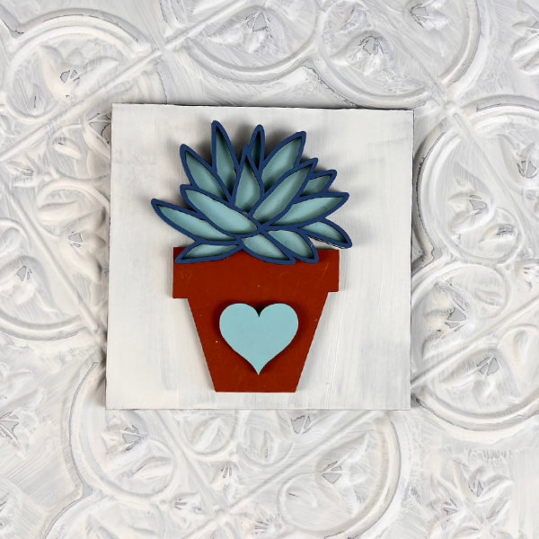 Introducing the Home Family Interchangeable Squares from Janet's Craft Corner: a beautifully decorative wooden piece that's perfect for home decor. It showcases a brown pot adorned with a heart symbol, gently cradling blue-green succulents set against a white textured background with intricate patterns. This charming item is part of our exclusive collection of interchangeable mini signs.