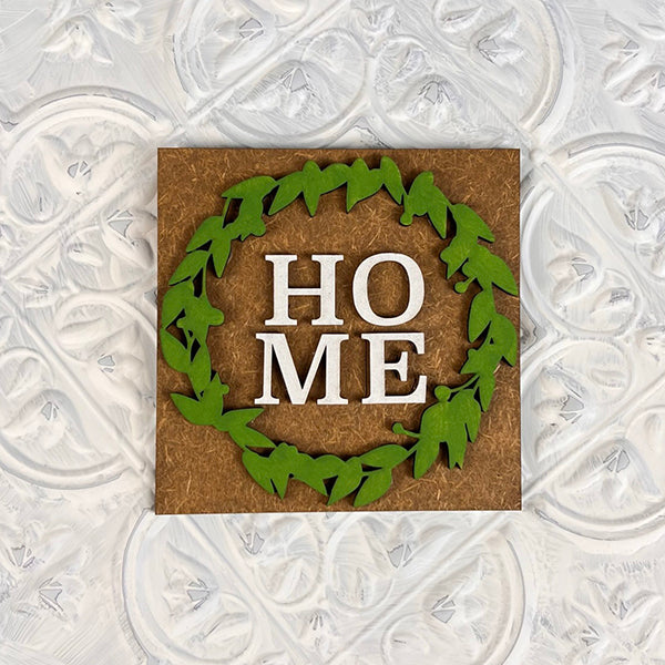 The "Home Family Interchangeable Squares" by Janet's Craft Corner features a square mini sign with the word "HOME" split over two lines. It is surrounded by a green leafy wreath against a brown background, making it perfect for home decor. This versatile piece can be displayed on a textured white surface.