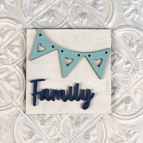 Introducing the Home Family Interchangeable Squares by Janet's Craft Corner: This charming decor piece boasts a blue pennant banner adorned with heart cutouts and the word "Family" beautifully scripted in blue cursive letters on a textured white background, making it the ideal addition to enhance your home decor.