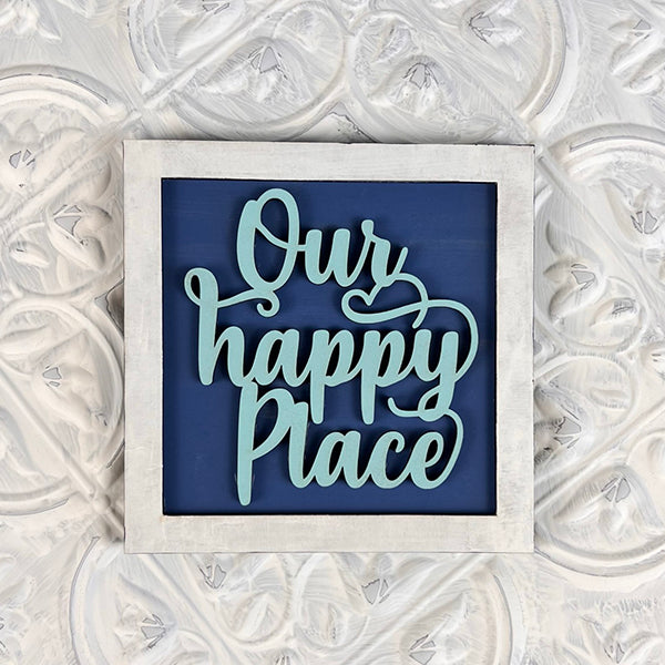 The "Home Family Interchangeable Squares" by Janet's Craft Corner features a framed sign with the phrase "Our Happy Place" in light blue cursive script on a dark blue background, bringing a charming element to your home decor. Its light-colored frame beautifully complements textured white decorative walls, making it an ideal choice for versatile signage in any environment.