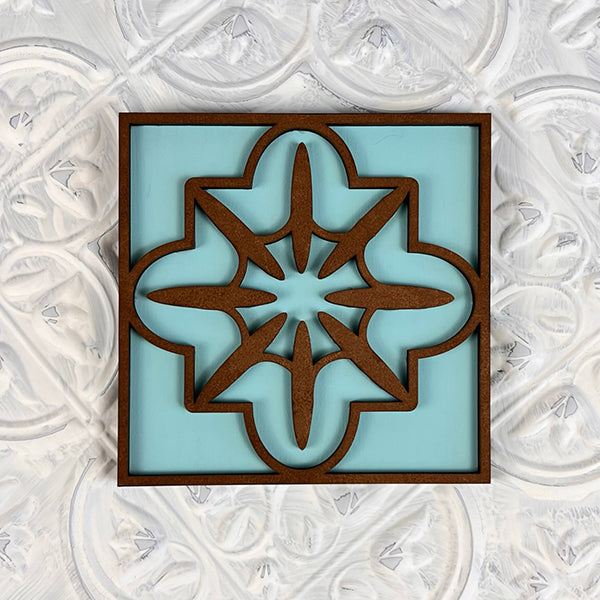 Introducing the Home Family Interchangeable Squares by Janet's Craft Corner: an exquisite wooden wall decor piece showcasing a symmetrical geometric design in dark brown set against a light blue background, resembling a star nestled within a floral shape. Ideal for enhancing any living space, this piece is elegantly mounted on a textured white wall for an added touch of sophistication.