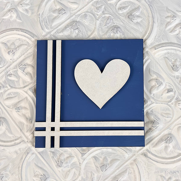 This eye-catching home decor item from Janet's Craft Corner, part of the "Home Family Interchangeable Squares" collection, showcases a square, blue design centered around a white heart. It features three parallel white lines in the lower left corner against a textured white background, perfect for fans of interchangeable signs in their living spaces.