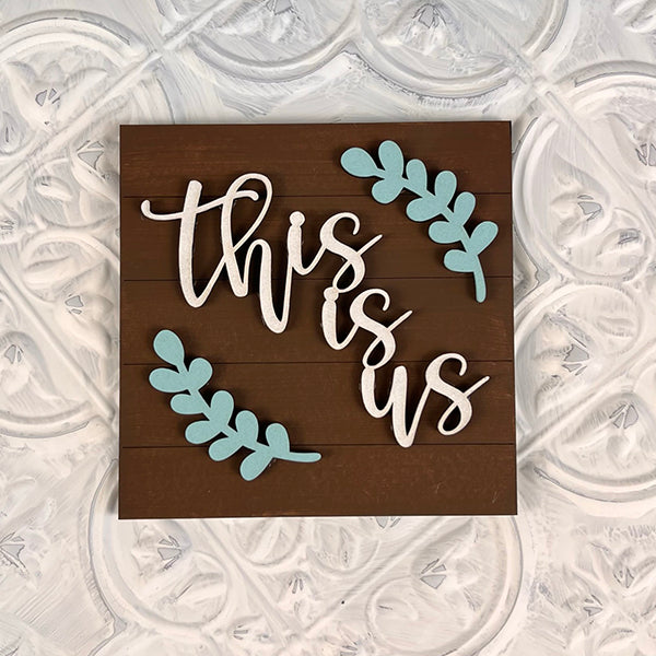 The Home Family Interchangeable Squares by Janet's Craft Corner is a delightful mini sign displaying the phrase "this is us" in elegant white cursive, complemented by three blue leaf designs. The background features a swirl pattern in calming white and beige tones, making it an ideal addition to your home decor.