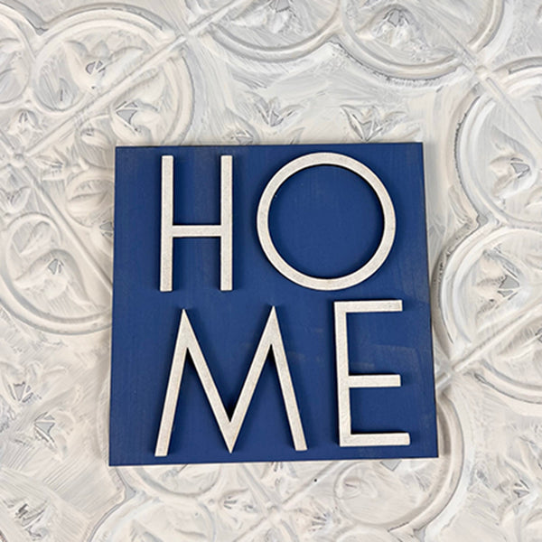 A decorative mini sign from Janet's Craft Corner, part of the "Home Family Interchangeable Squares" collection, features the word "HOME" in white letters on a square blue background against a textured white wall, adding a charming touch to your home decor.