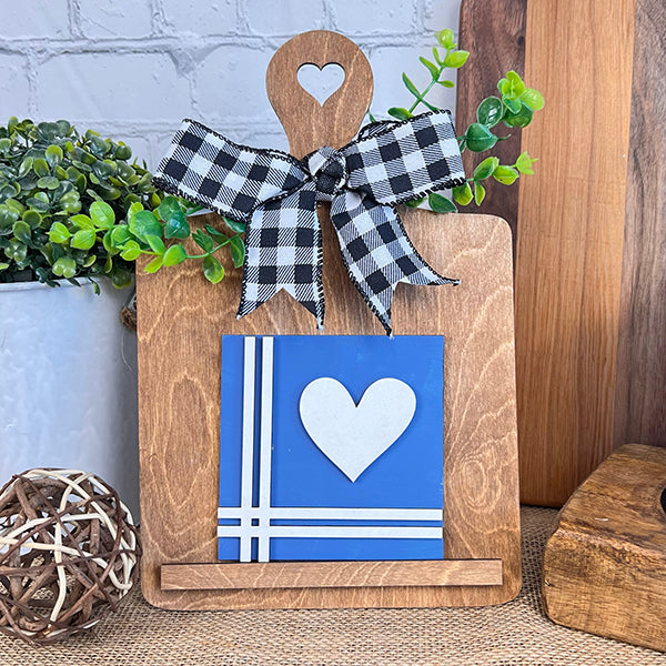 The Home Family Interchangeable Squares by Janet's Craft Corner is a delightful piece of home decor. This charming wooden cutting board features a heart-shaped cutout at the top, accentuated by a black and white checkered bow. At the center of the board is a blue square with a white heart and stripes. Greenery enhances its aesthetic appeal, while a wicker ball and mini signs complete this enchanting decoration.
