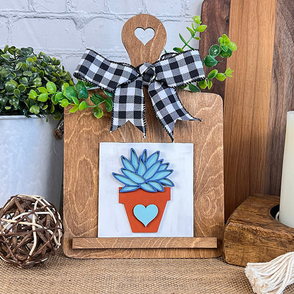 The "Home Family Interchangeable Squares" by Janet's Craft Corner is a delightful wooden stand featuring a heart cut-out at the top and decorated with a black and white checkered ribbon. Perfect for home decor, it showcases a design with potted succulents. A charming arrangement of green plants, a woven ball, and a candle complement this piece beautifully.