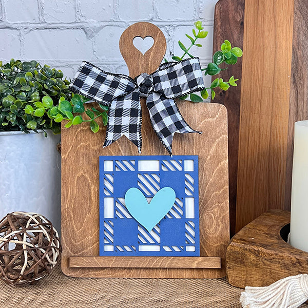 Crafted by Janet's Craft Corner, the Home Family Interchangeable Squares are a delightful piece of home decor. This wooden stand is designed with a charming heart cutout at the top and is embellished with a black and white checkered ribbon. It features greenery emerging from behind a blue and white geometric square showcasing a blue heart in the center, making it an ideal addition to your collection of interchangeable mini signs on any tabletop.