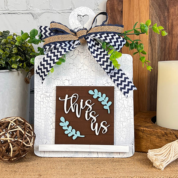 Product: Home Family Interchangeable Squares by Janet's Craft Corner

Description: This charming piece features "This is us" elegantly written in white cursive on a natural wooden background, enhanced with blue leaf accents. It is mounted on a shabby chic white board and embellished with a stylish black-and-white chevron ribbon accompanied by sprigs of green leaves. Perfect for enhancing your home decor when placed amongst candles and plants.