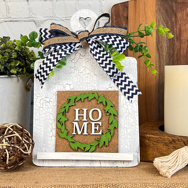 This delightful home decor item, the "Home Family Interchangeable Squares" from Janet's Craft Corner, showcases a white stand beautifully adorned with a green leaf wreath encircling the word "HOME." It features decorative elements like a black and white chevron ribbon, greenery, and a wicker ball. Additionally, it comes with mini signs that allow you to effortlessly create an inviting and customizable cozy atmosphere.