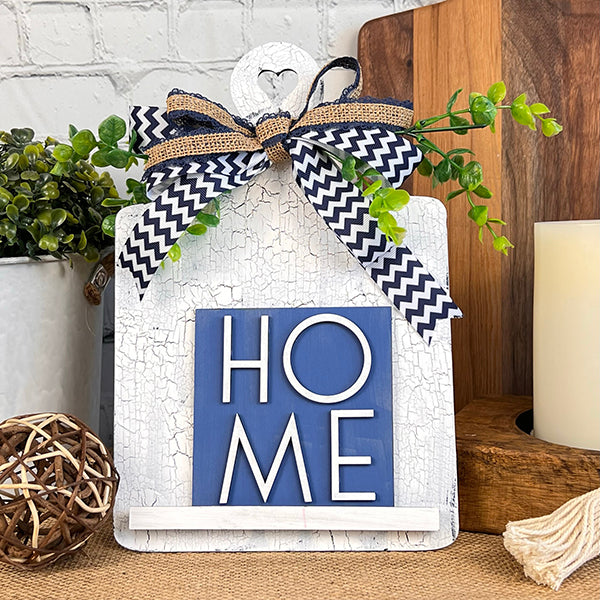 Janet's Craft Corner presents the "Home Family Interchangeable Squares," a delightful piece of home decor. This decorative wooden plaque, featuring a heart-shaped top and the word "HOME" in white on a blue background, is enhanced with a chevron-patterned bow and greenery. It's beautifully set against a rustic kitchen backdrop with plants and candles, making it ideal for mini sign enthusiasts.