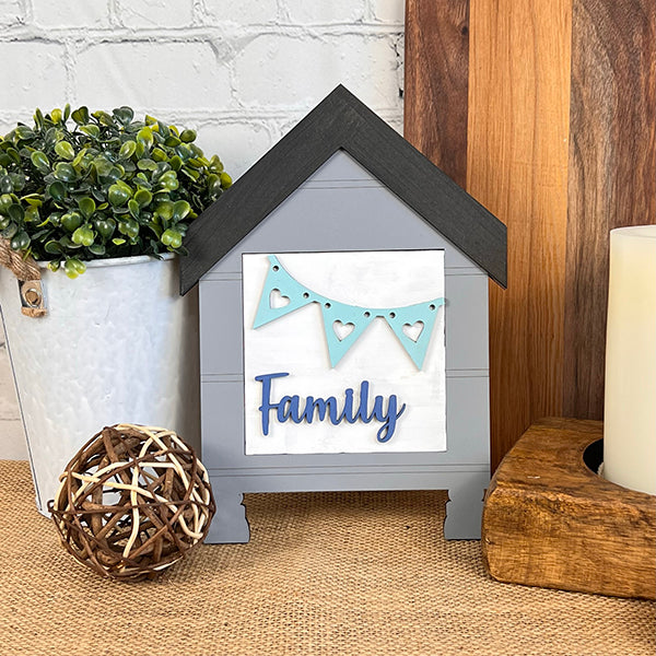 The Home Family Interchangeable Squares by Janet's Craft Corner is a charming wooden house-shaped sign featuring the word "Family" and embellished with a teal heart-accented banner. It's an ideal piece of home decor, beautifully positioned among a wicker ball, candle, potted plant, and wooden cutting board against a brick wall.