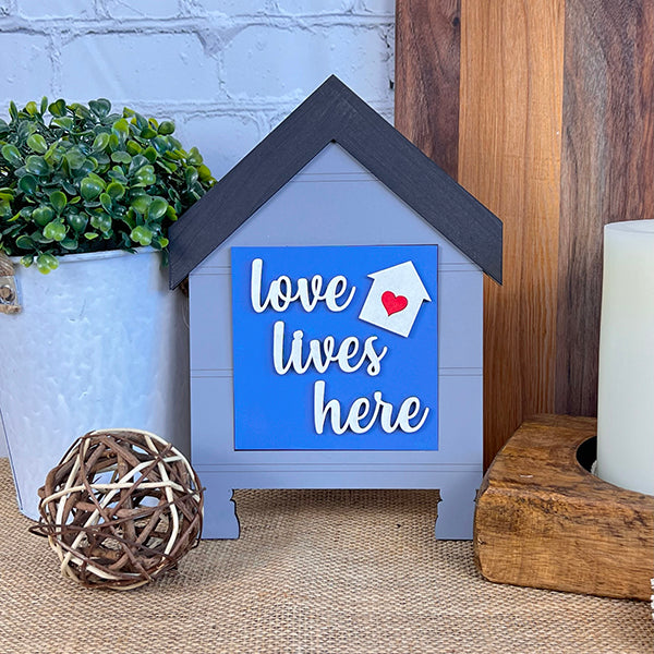 The Home Family Interchangeable Squares by Janet's Craft Corner is a delightful mini sign shaped like a house featuring a black roof and the phrase "love lives here" on a blue background. Ideal for home decor, it includes an endearing red heart, and when displayed alongside a potted plant, wicker ball, and large candle, it creates a charming scene against a white brick wall.